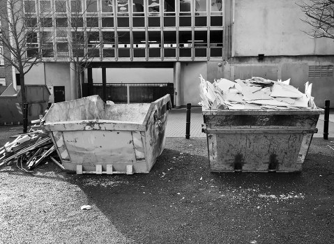 In an outdoor urban area, two large industrial-sized skip bins are positioned on a paved surface in front of a modern building with a grid-like facade. The left skip is filled with large, broken construction debris, such as concrete slabs and broken wooden panels, showing rough, uneven textures and light grey to beige tones. The right skip is filled with crumpled, layered white and light grey waste materials, possibly paper or cardboard. Both skips are made of metal, with visible signs of rust and dirt, indicating frequent use. Small debris and scattered particles are visible on the ground around the skips, and a black safety bollard sits behind them. In the background, the building's concrete wall and a ground-level garage or loading area are visible, with part of the pavement and a few leafless trees framing the scene. The overall setting suggests a waste collection or rubbish removal operation tied to commercial or office clearance services in Stoke Newington, as referenced by Office Clearance Stoke Newington.