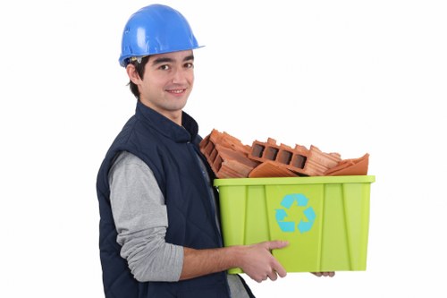 A young man wearing a blue safety helmet and navy blue work vest over a grey long-sleeve shirt holds a green plastic recycling bin with a blue recycling symbol on its side. The bin contains a stack of red clay bricks, neatly arranged with some edges visible, indicating the disposal of construction waste. The background is plain white, emphasizing the focus on the individual and the debris he is holding. This scene relates to rubbish removal services provided by Office Clearance Stoke Newington, situated in the Stoke Newington area. The bricks' rough texture and reddish-brown color contrast with the smooth, vibrant surface of the recycling bin, highlighting the type of waste managed in local clearance projects. The individual’s friendly expression and professional attire reflect a responsible approach towards waste collection and recycling within the community, suitable for website imagery aimed at potential clients seeking efficient rubbish removal solutions in London’s north-eastern boroughs.
