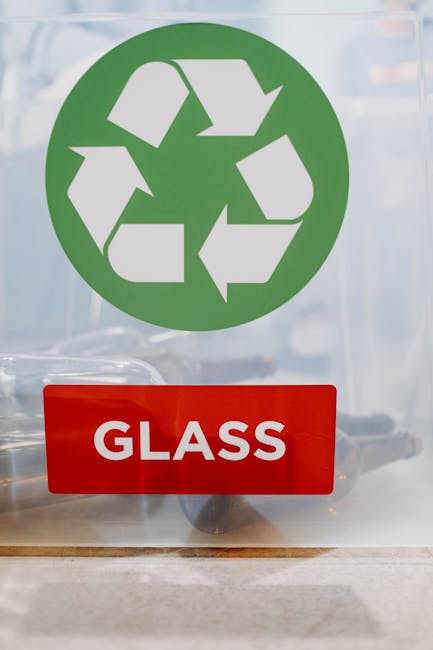 A clear glass door or panel displaying a large green recycling symbol composed of three arrows forming a triangle, indicating recyclable materials. Below, there is a red rectangular sign with bold whi