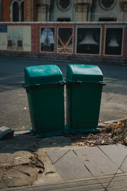 Two upright green plastic rubbish bins with closed, slightly domed lids are positioned on a concrete sidewalk near the edge of a street. The bins are made of sturdy textured plastic with visible hinge