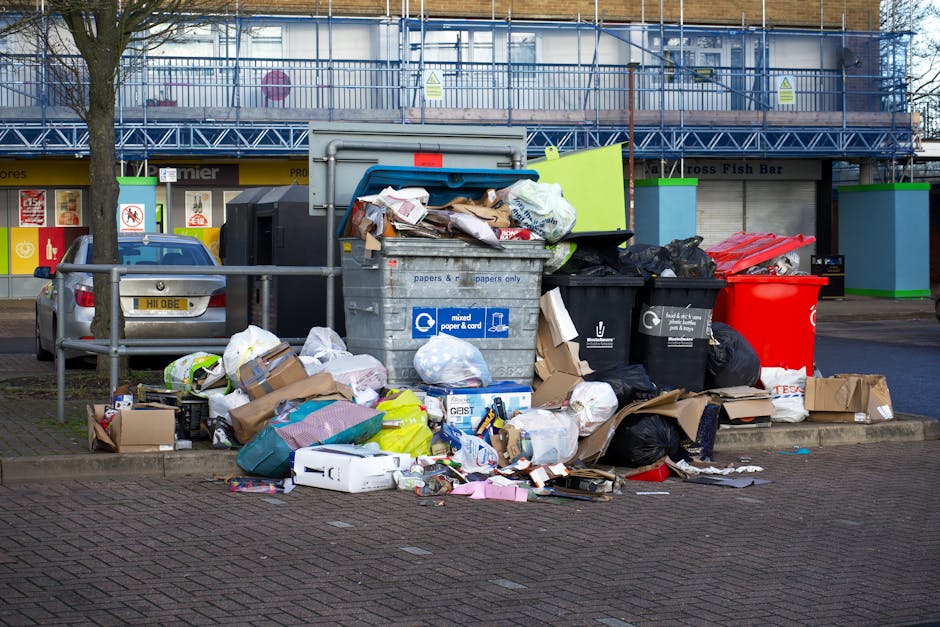 A scene of a large pile of mixed waste materials and discarded items outdoors on a rough, uneven surface, with various bags of rubbish, broken furniture, and miscellaneous debris scattered across the