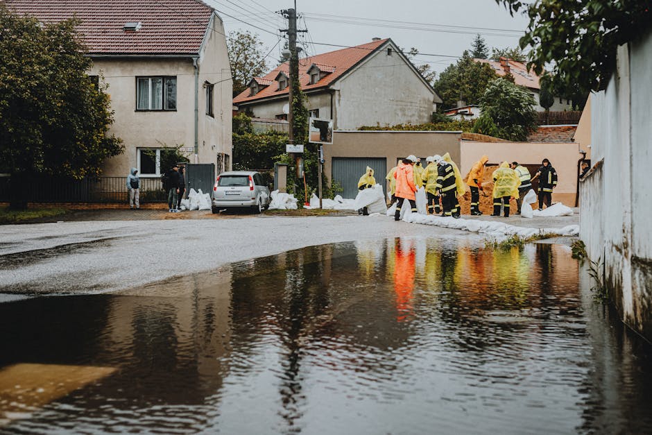 A group of emergency rescue workers walking through a flooded street, wearing high-visibility yellow vests, orange and black waterproof gear, and helmets, with some carrying rescue equipment such as w