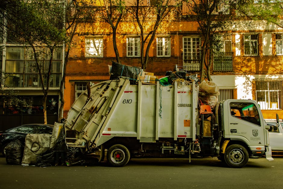 A small burgundy flatbed truck parked on a city street in front of a modern office building with large glass windows and a concrete facade. The truck's cargo bed is filled with various types of waste