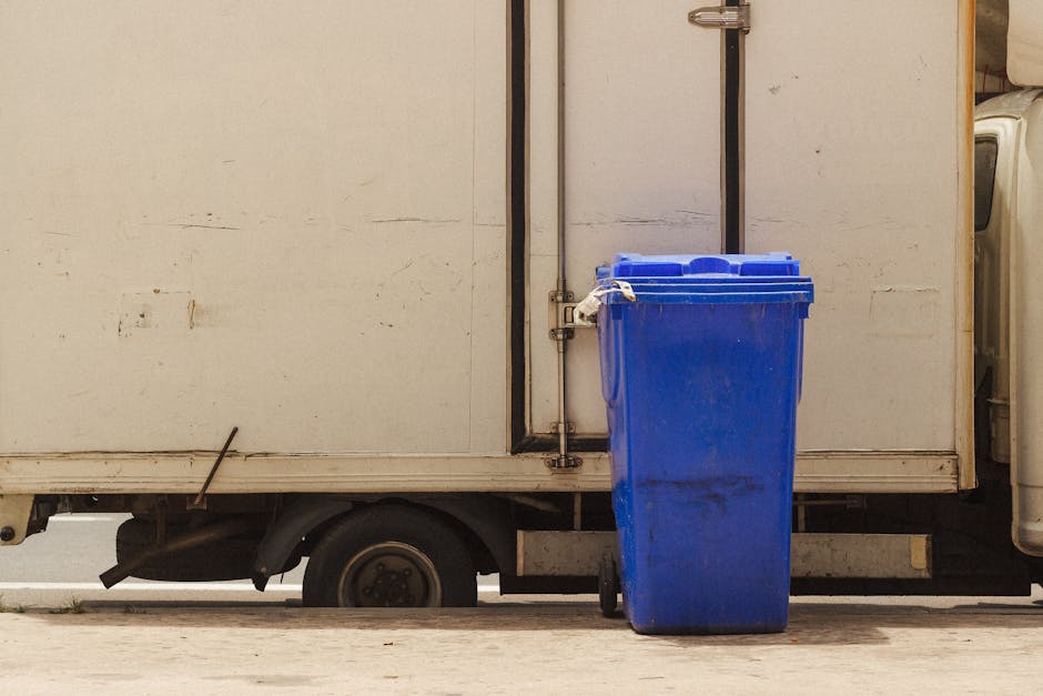 A bright blue plastic wheelie bin with a closed lid is positioned on the pavement next to a white delivery truck or storage unit. The bin's surface appears slightly scuffed with some grime near the bo